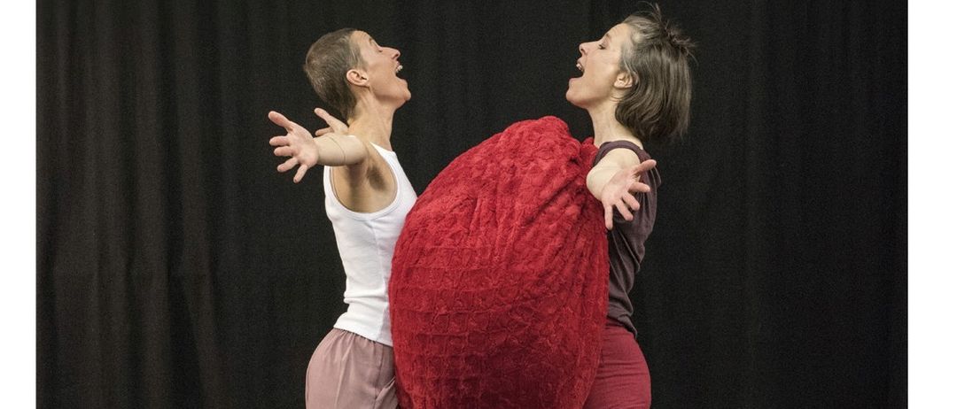 Zwei Frauen führen eine Tanzroutine auf einer Bühne auf, wobei ein rotes herzförmiges Luftballon in der Nähe schwebt. Das Datum und die Uhrzeit der Veranstaltung sind in der oberen linken Ecke angezeigt.