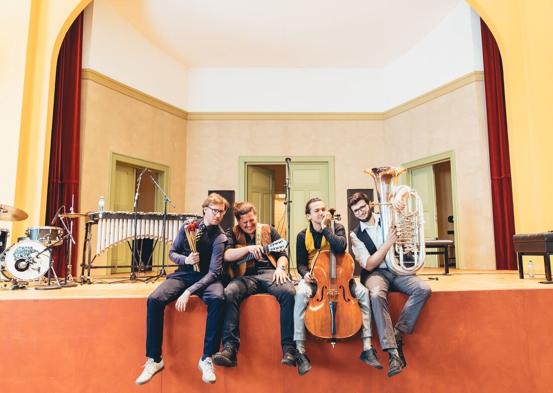 Fünf Männer sitzen auf einer Bühne und lächeln für ein Foto. Sie halten Musikinstrumente, darunter ein Tuba, Cello, Gitarre und Xylophon. Die Bühne hat einen Holzboden und ist von Türen umgeben.