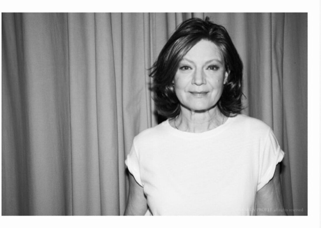 Ein Schwarz-Weiß-Porträt einer Frau mit kurzen Haaren und einem weißen T-Shirt, das vor einem Vorhang steht.