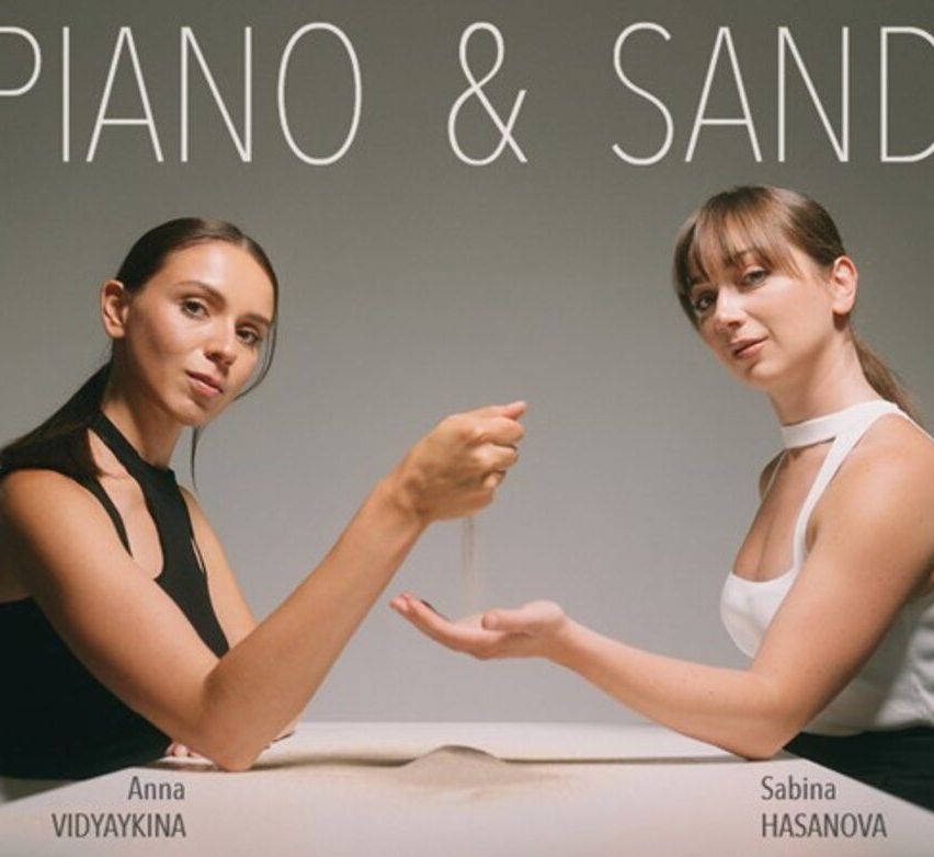 Zwei Frauen sitzen an einem Tisch, Blickrichtung gegeneinander. Eine hält eine kleine Menge Sand. Der Text darüber lautet 'Piano & Sand'. Die Namen unten lauten 'Anna Vidyakina' und 'Sabina Hasanova'.