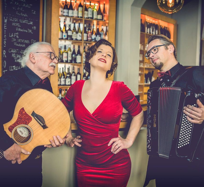 Drei Musiker in einer Bar: eine Frau in einem roten Kleid, ein Mann mit Brille, der eine Gitarre hält, und ein Mann, der Akkordeon spielt.