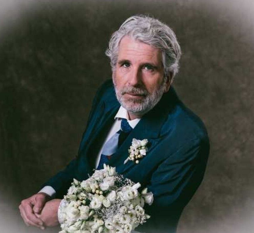 Ein Mann mit grauem Haar und Bart trägt einen dunkelblauen Anzug, ein weißes Hemd und eine Krawatte. Er hält einen Strauß weißer Blumen.