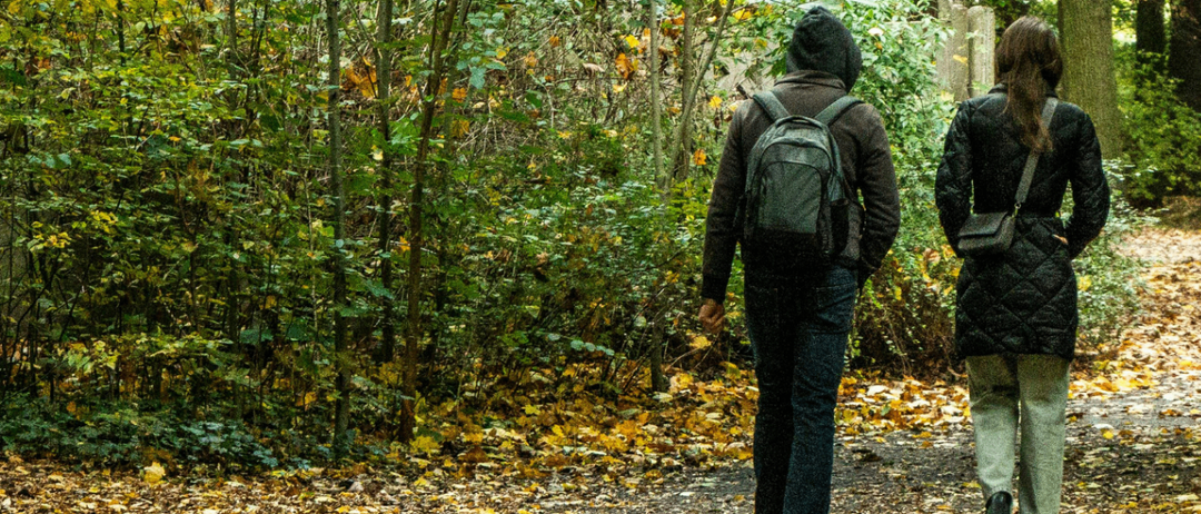 Two people walk on a path in a forest, surrounded by trees with autumn leaves. One wears a backpack and a beanie, the other a quilted jacket and sneakers.