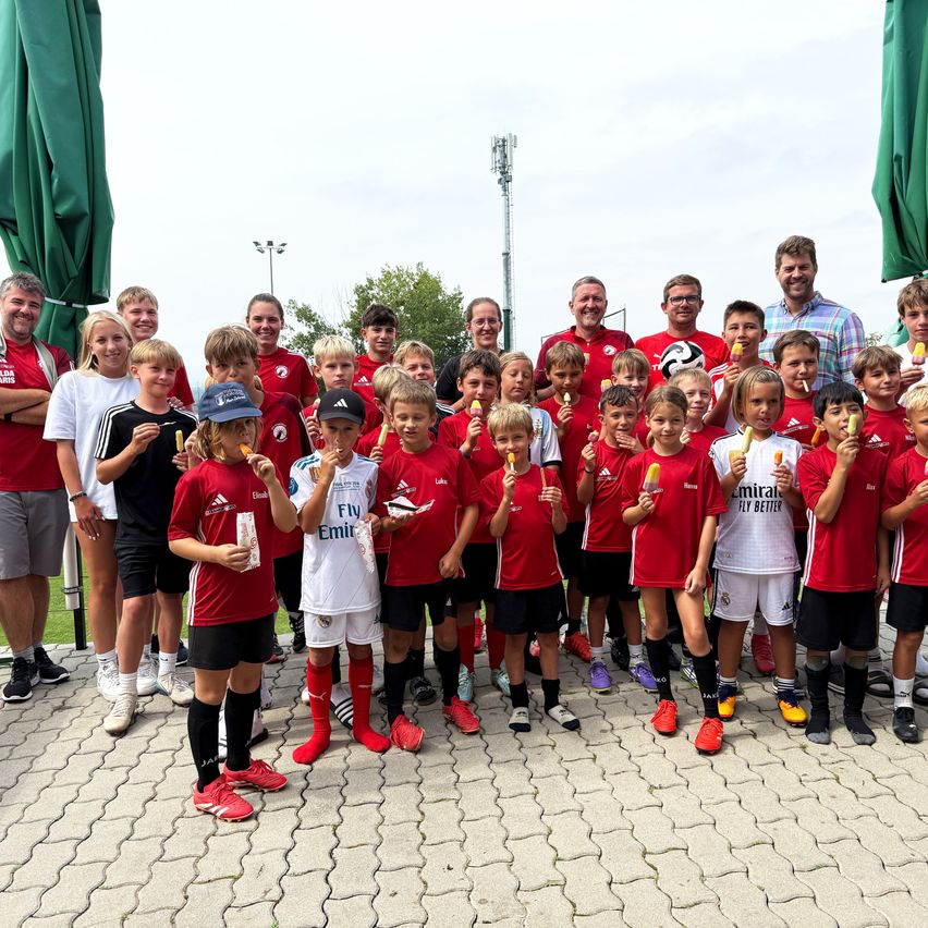 Eine Gruppe von Kindern und Erwachsenen, gekleidet in roten und weißen Sporttrikots, posiert für ein Foto und hält Eispops. Hinter ihnen stehen grüne Regenschirme und Bäume.