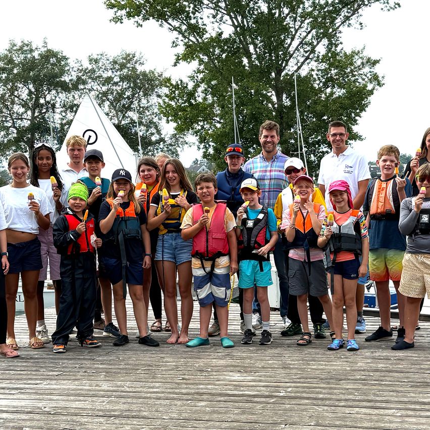 Eine Gruppe von Kindern und Erwachsenen steht auf einem Steg in Schwimmwesten, einige halten Eislutscher. Sie lächeln für ein Foto, mit Booten und Bäumen im Hintergrund.