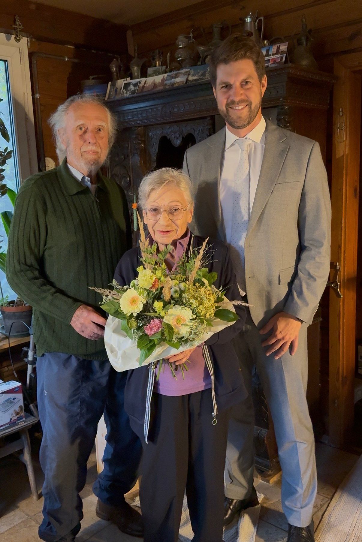 Three people stand together, smiling. The woman holds a colorful bouquet of flowers. The man beside her wears glasses and a green sweater. The man on the right wears a gray suit and tie. Behind them is a window with a plant.