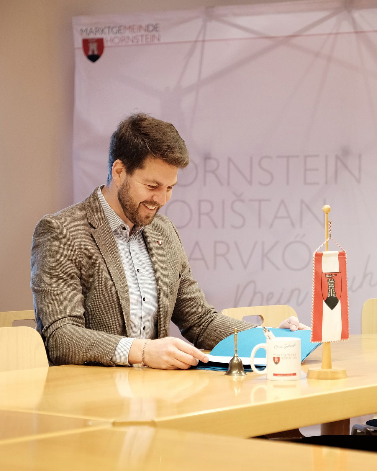 Ein Mann sitzt lächelnd an einem Tisch, mit einem Stift und einer Tasse. Hinter ihm sind eine Flagge und ein Banner zu sehen.