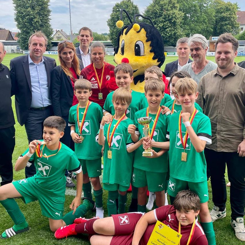 Eine Gruppe junger Fußballer in grünen Trikots posiert mit einem Bienenmaskottchen und Erwachsenen, hält Medaillen und einen Pokal.
