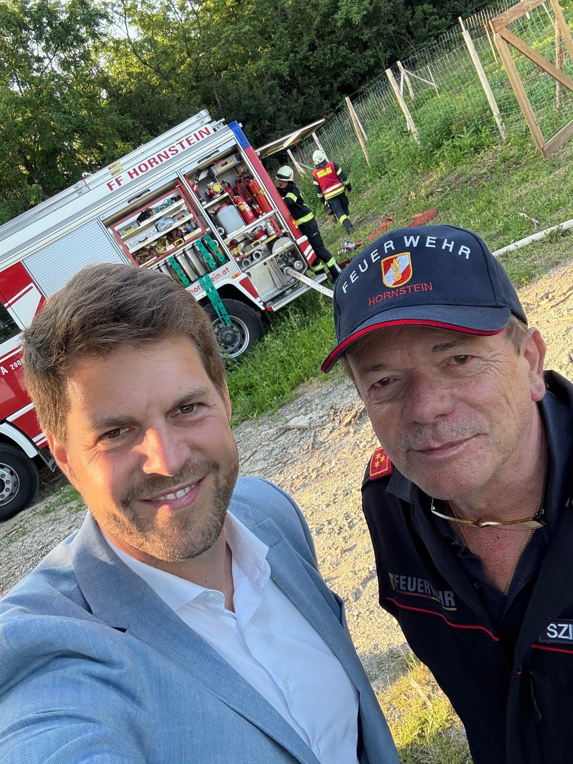 Zwei Männer lächeln für ein Selfie, mit einem Feuerwehrwagen im Hintergrund. Ein Mann trägt eine dunkelblaue Kappe mit 'Feuerwehr Hornstein'. Hinter ihnen arbeiten zwei Feuerwehrleute in der Nähe eines Feuerwehrwagens.
