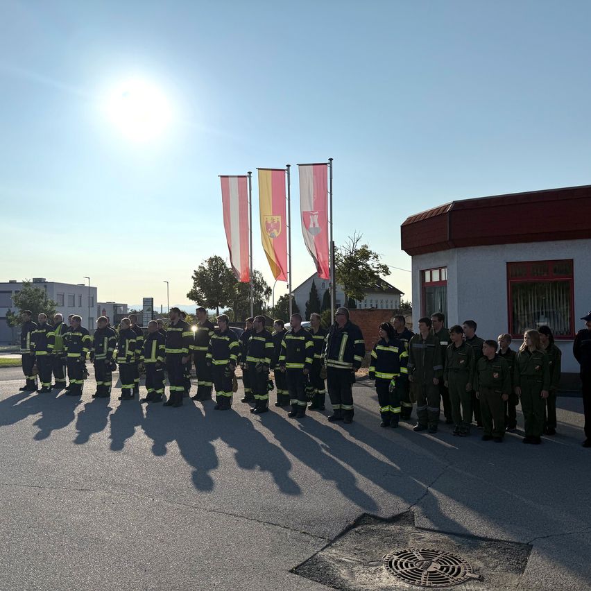 Eine Gruppe von Feuerwehrleuten, darunter auch Kinder, steht in einer Reihe mit Fahnen hinter ihnen an einem sonnigen Tag.