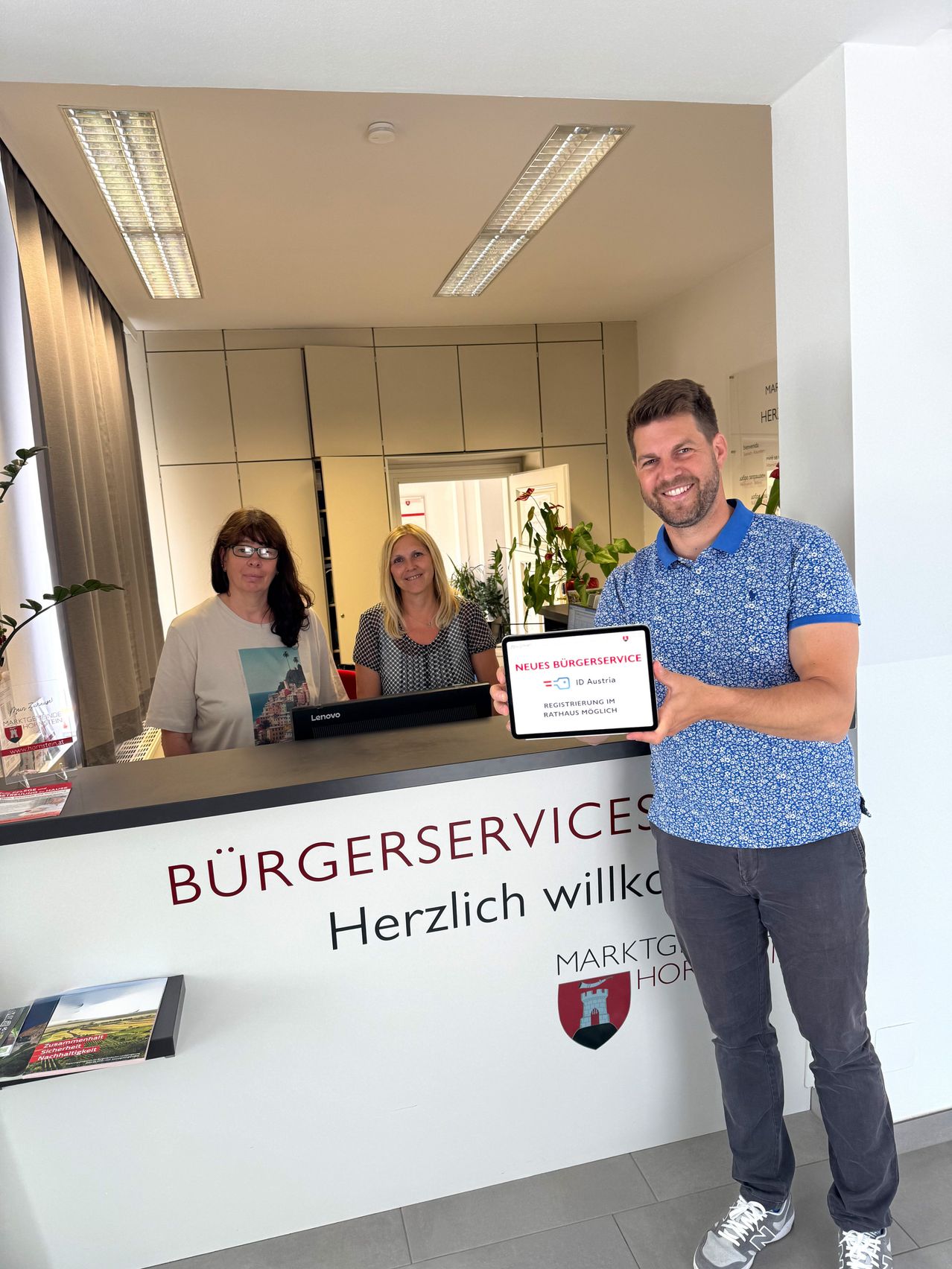 Ein Mann hält ein Zertifikat vor einer Rezeption. Zwei Frauen stehen hinter dem Tresen, eine hält einen Laptop. Das Schild steht Burgerservices Herzlich willkommen.