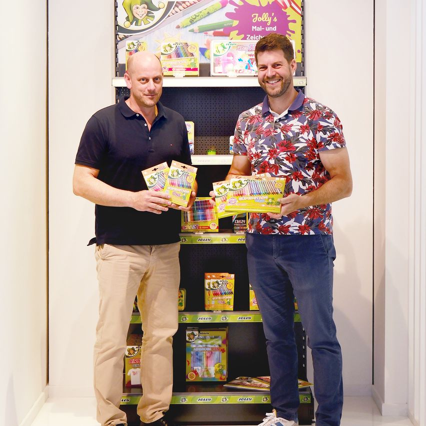 Zwei Männer stehen neben einem Regal voller Kunstmaterialien und lächeln. Sie halten Boxen mit Buntstiften in den Händen. Der Hintergrund zeigt eine bunte Produktpalette.