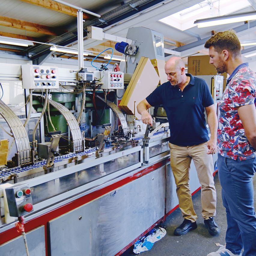 Zwei Männer in einer Fabrik, einer inspiziert eine Maschine, der andere beobachtet. Die Maschine hat mehrere Räder und Bedienfelde.