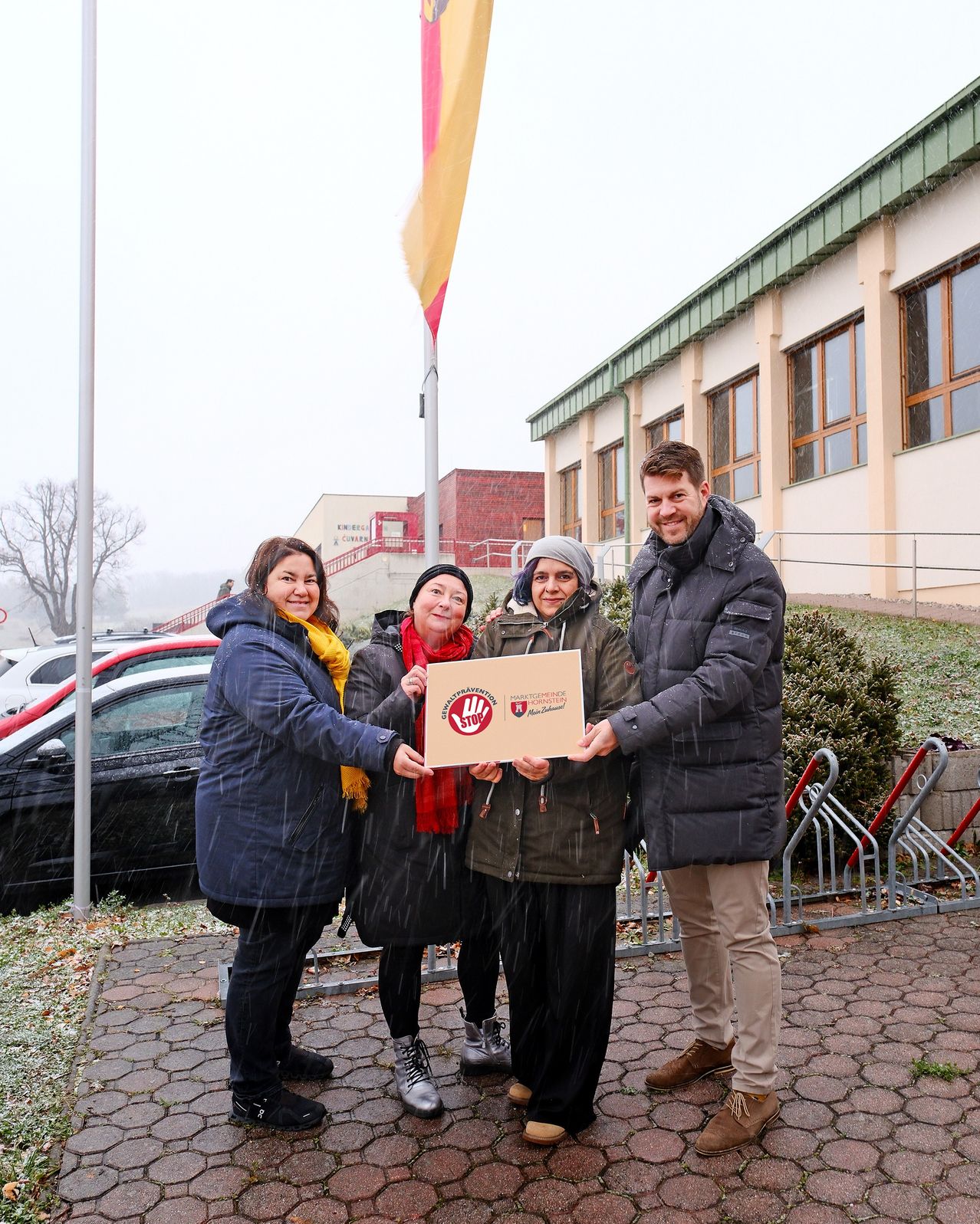 Vier Menschen stehen an einem nassen Tag draußen. Drei von ihnen halten ein Poster mit einem Logo und lächeln. Dahinter befindet sich ein Gebäude mit einer Flagge.
