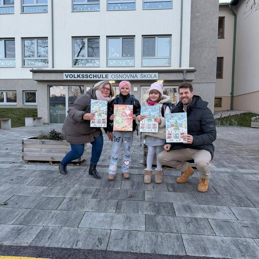 Vier Personen posieren für ein Foto vor einer Schule und halten Bücher in der Hand. Zwei Kinder stehen zwischen zwei Erwachsenen. Das Schulschild lautet Volksschule Osnovna skola.