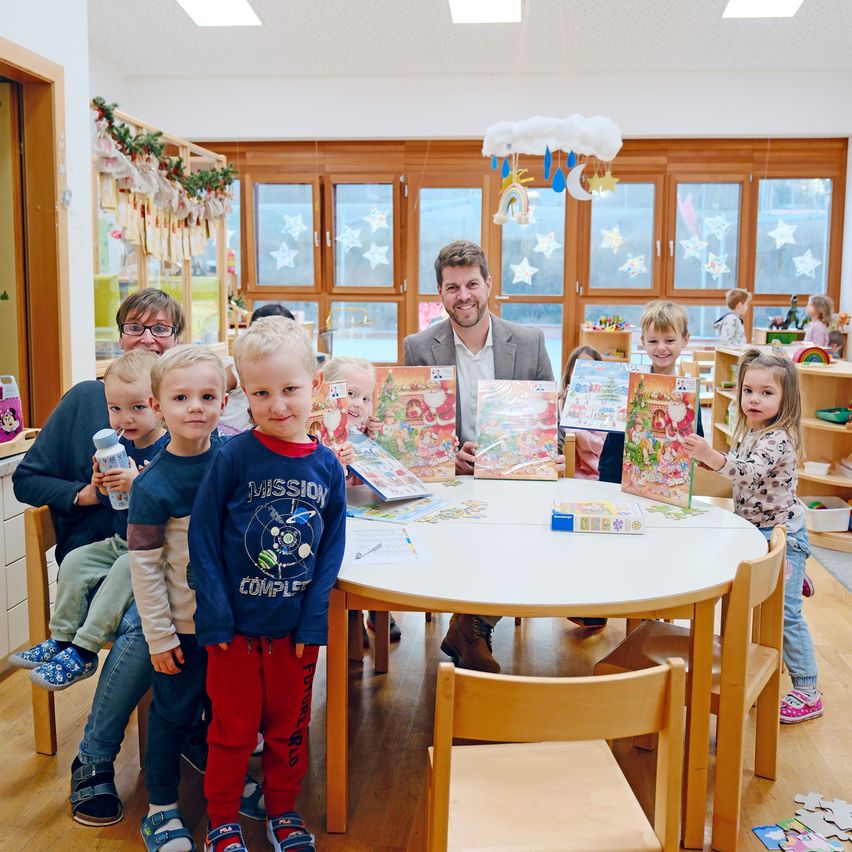Eine Gruppe von Kindern, Erwachsenen und einem Mann befindet sich in einem Raum mit Holzboden und Fenstern. Sie lächeln und halten weihnachtliche Bücher und Puzzles in den Händen.