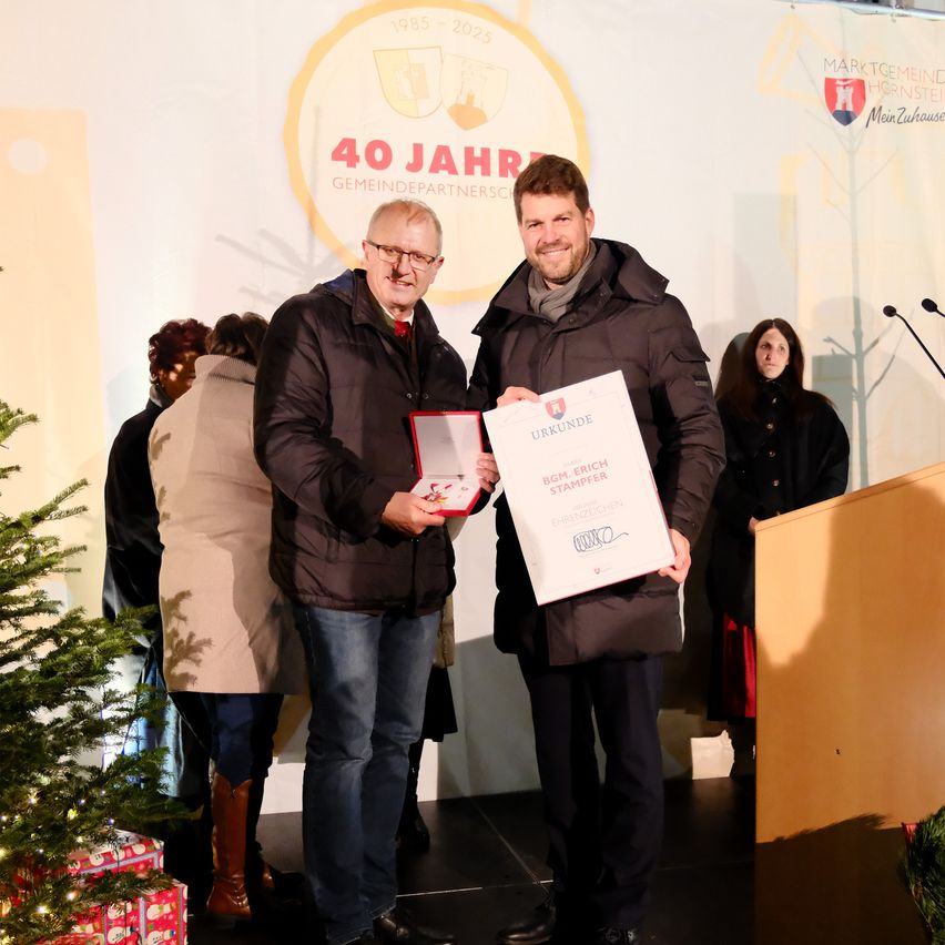 Zwei Männer stehen auf einer Bühne, einer hält eine Urkunde und lächelt für ein Foto. Hinter ihnen stehen andere, mit einem Weihnachtsbaum links und einem Podium rechts. Im Hintergrund steht '40 Jahre Gemeindepartnerschaft'.