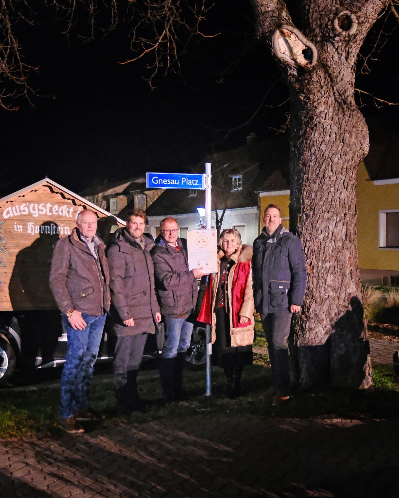 Fünf Personen stehen vor einem Straßenschild für Gneenau Platz, eine hält ein Zertifikat. Sie tragen Winterkleidung. Hinter ihnen steht ein hölzerner Wagen mit den Worten 'ausgysteckt in Bornheim'.