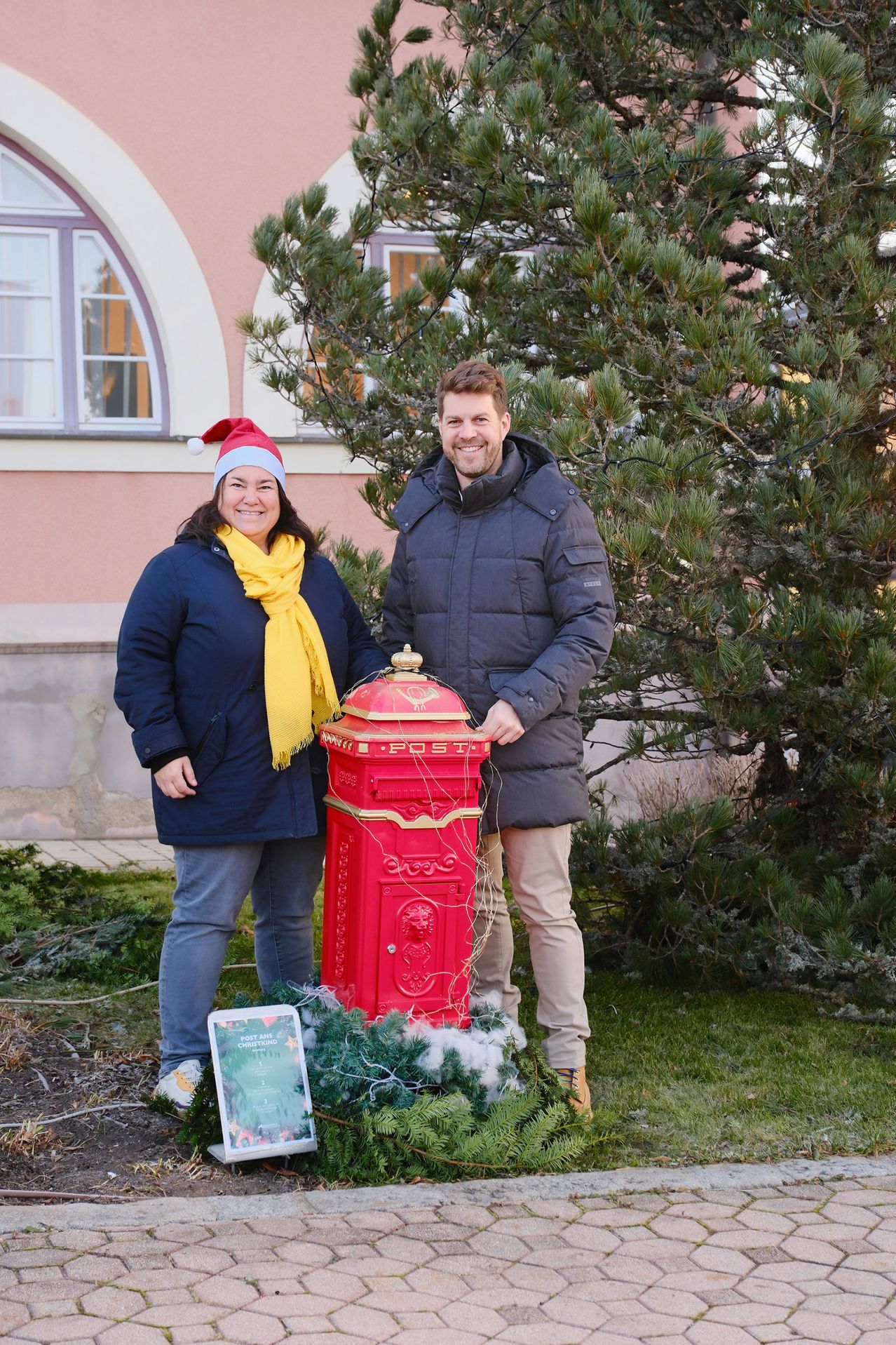 Ein Mann und eine Frau stehen vor einem roten Briefkasten, der mit Weihnachtsdekorationen geschmückt ist. Sie tragen beide Winterkleidung, und die Frau hat einen Weihnachtsmannhut und ein gelbes Tuch um den Hals. Sie lächeln und scheinen für ein Foto zu posieren.