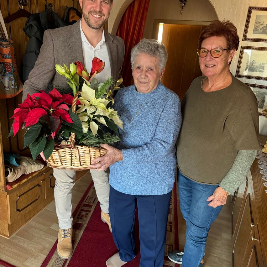 Drei Personen posieren drinnen mit einer Blumendekoration. Ein Mann in einem Anzug hält einen Korb mit Blumen. Zwei Frauen stehen neben ihm, eine in einem blauen Pullover und die andere in einem grünen Oberteil.