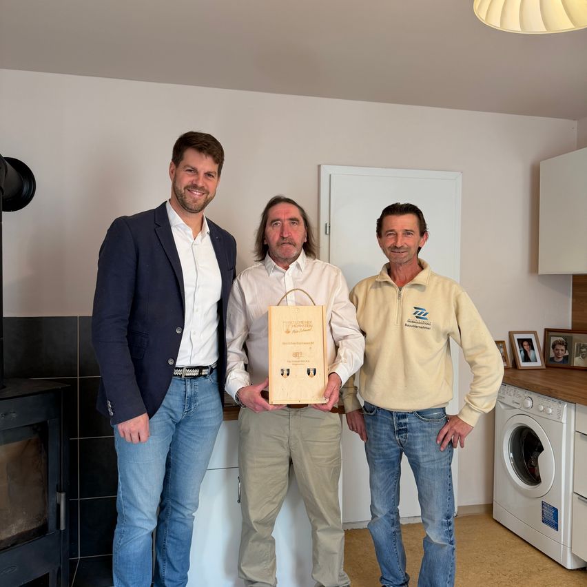 Drei Männer stehen in einer Küche, einer hält eine Box. Hinter ihnen ist eine weiße Tür und Schränke zu sehen. Ein Kamin und eine Waschmaschine befinden sich auf beiden Seiten.