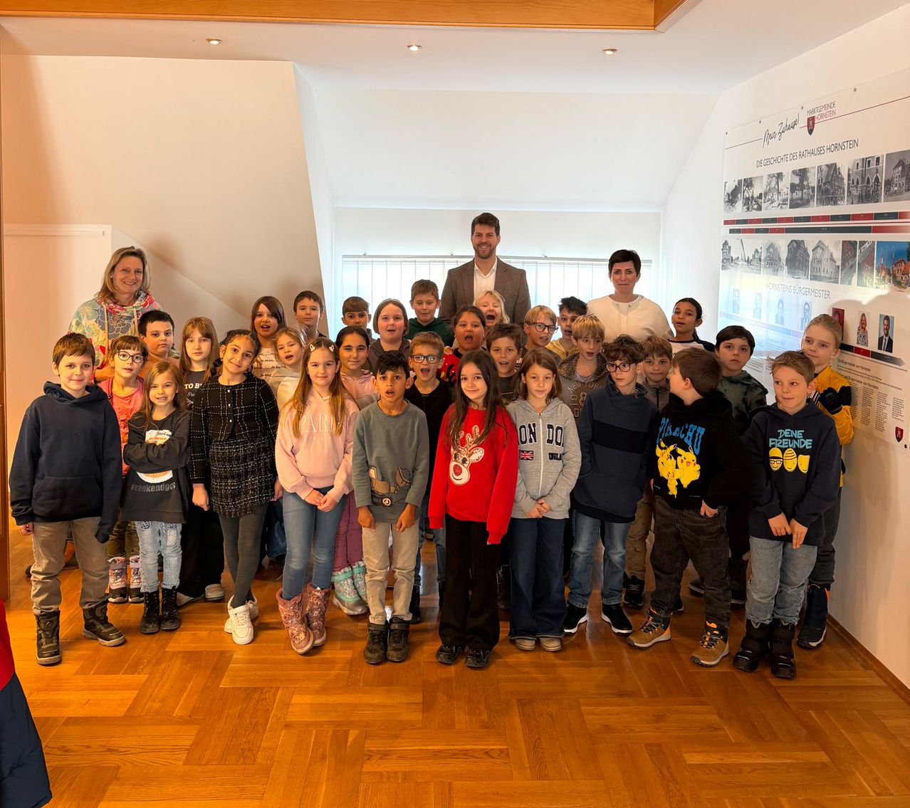 Eine Gruppe von Kindern und zwei Erwachsenen stehen auf einem Holzboden und posieren für ein Foto. Ein Mann in einem Anzug steht in der Mitte. Eine Wand mit Bildern und Text steht dahinter.
