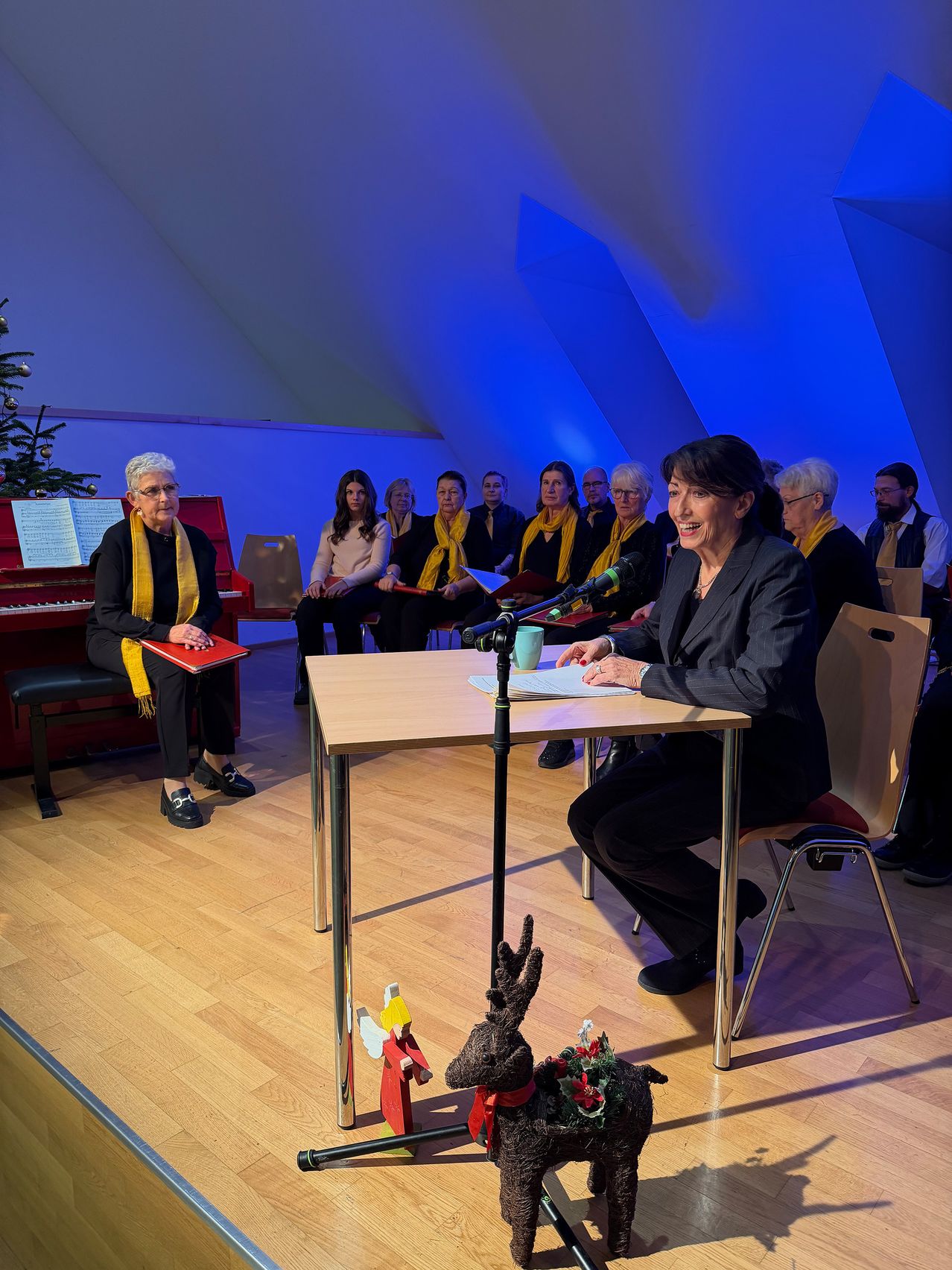 Eine Frau spricht in ein Mikrofon an einem Tisch. Hinter ihr sitzt ein Chor von Frauen in gelben Schals auf Stühlen. Eine Frau hält eine rote Mappe. Links steht ein Weihnachtsbaum.