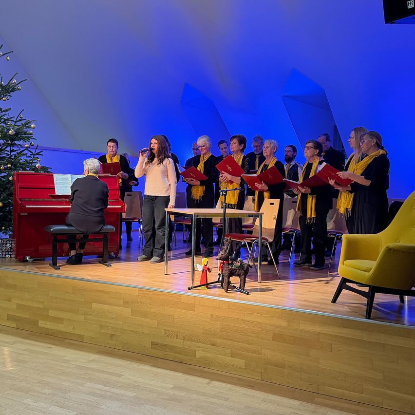Ein Chor tritt auf einer Bühne mit einem Pianisten, einem Weihnachtsbaum und Zuhörern auf. Die Chormitglieder tragen Schals und der Pianist sitzt an einem roten Klavier.