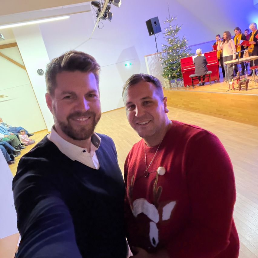 Zwei Männer posieren für ein Foto in einem Raum mit einem Weihnachtsbaum und einem Chor im Hintergrund.