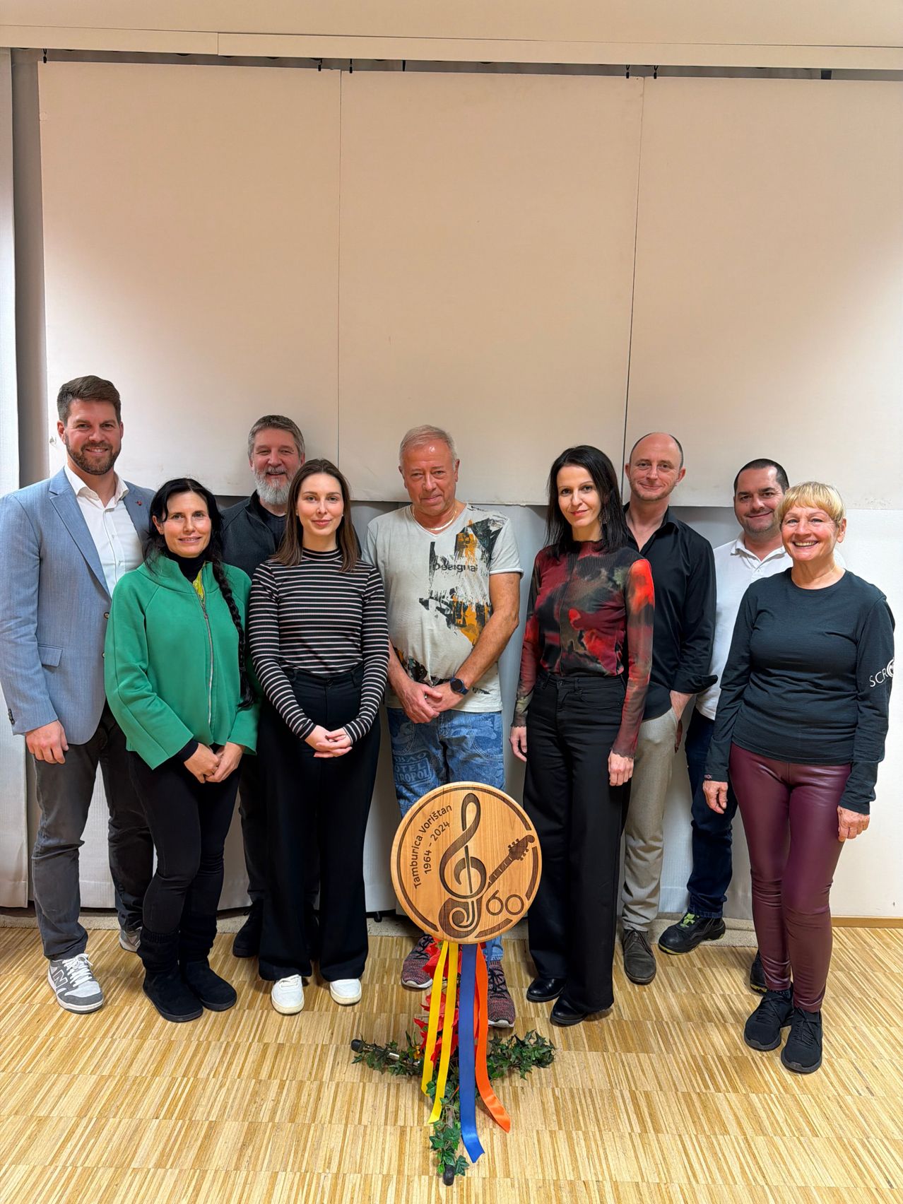 Eine Gruppe von Menschen, einschließlich Männer und Frauen, steht lächelnd vor einer Wand mit einer Holzplakette. Die Plakette hat ein musikalisches Notendesign mit den Worten 'Tamburitza-Festival 1964-2024' darauf. Sie nehmen wahrscheinlich an einer Veranstaltung teil.