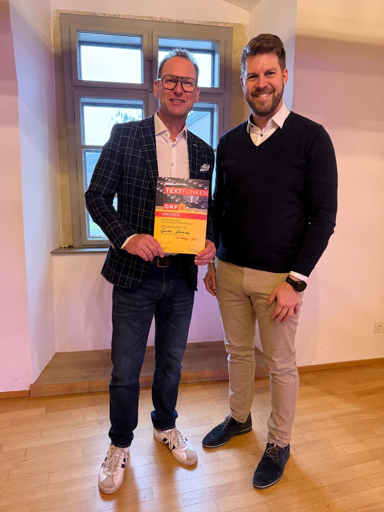 Zwei Männer stehen drinnen neben einem Fenster. Der Mann auf der linken Seite trägt einen karierten Blazer und Jeans und hält eine Urkunde. Der Mann auf der rechten Seite trägt einen dunklen Pullover und Khaki-Hosen und lächelt.