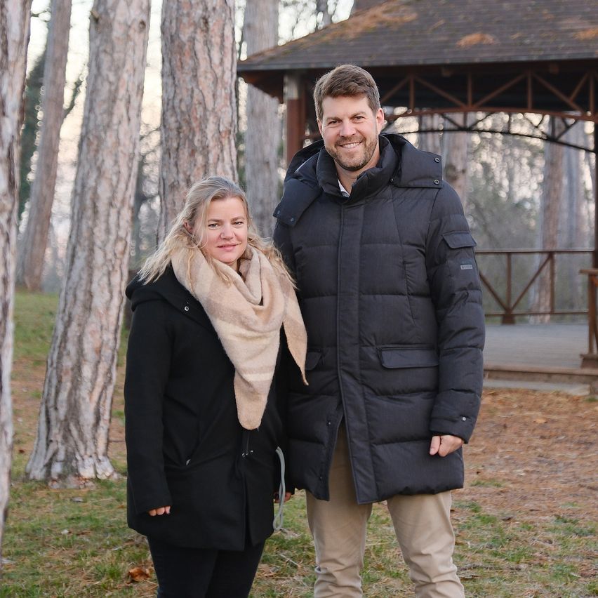 Ein Mann und eine Frau stehen gemeinsam in einem Park. Sie lächeln und scheinen für ein Foto zu posieren. Die Frau trägt einen Schal und einen schwarzen Mantel, während der Mann einen schwarzen Jacken trägt. Hinter ihnen steht ein hölzerner Pavillon mit Dach. Das Gebiet ist von hohen Bäumen umgeben.