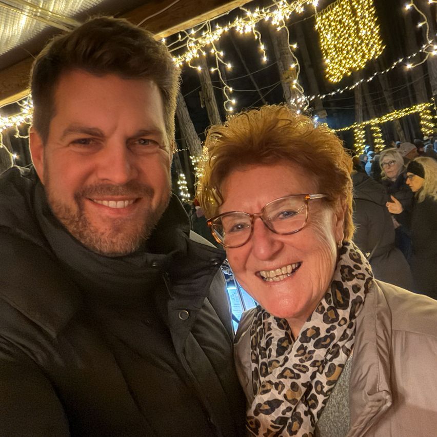 Ein lächelndes Paar steht eng beieinander, warm gekleidet, in einer festlichen Umgebung mit Lichterketten und einem verschwommenen Hintergrund anderer Menschen.