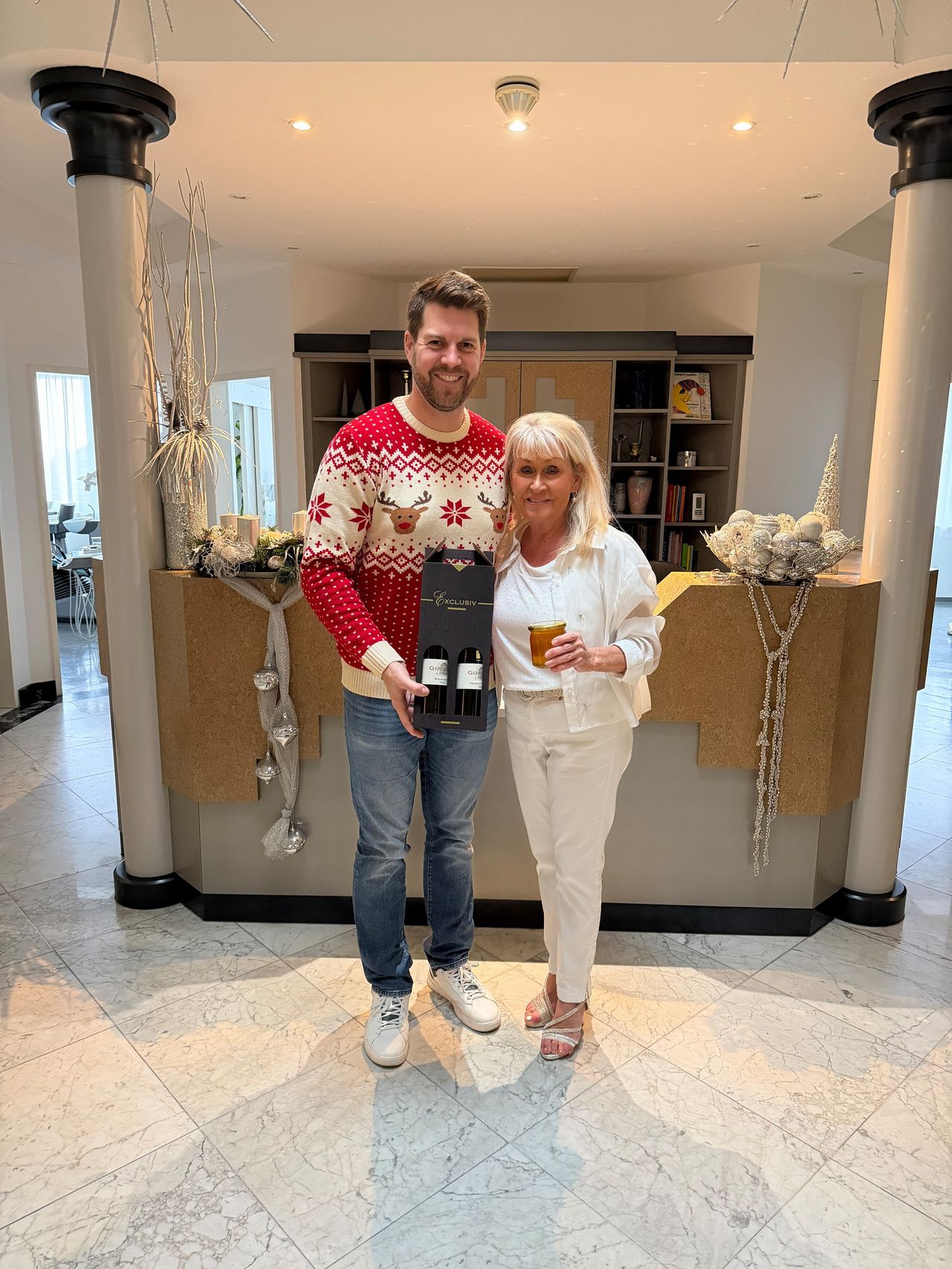 Ein Mann und eine Frau stehen in einem Raum mit festlichen Dekorationen. Der Mann trägt einen Weihnachtspullover und hält eine Weinbox, während die Frau ein Glas hält. Sie lächeln beide. Dahinter befindet sich eine Theke mit Regalen und Ornamenten.