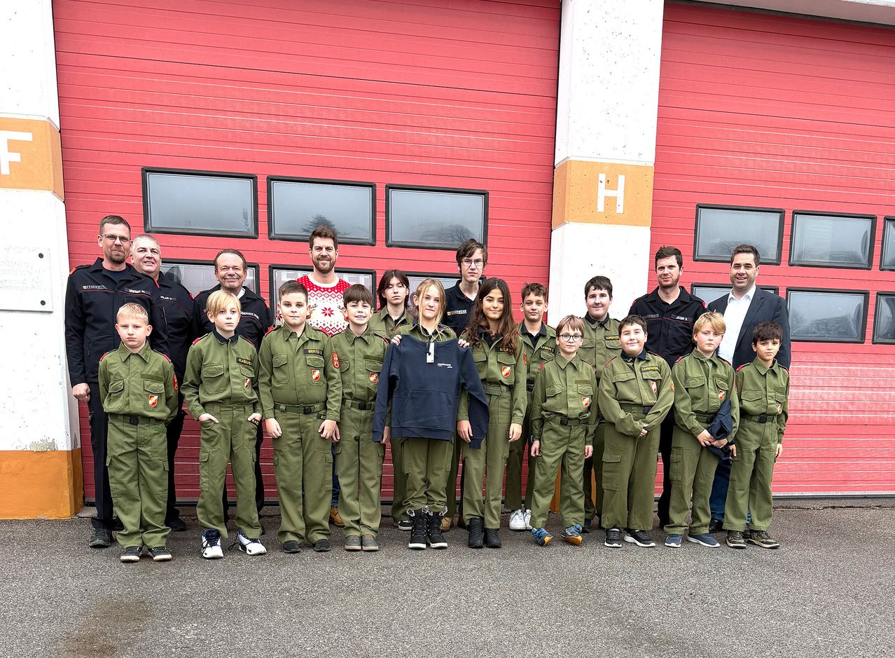 Eine Gruppe von Kindern in grünen Uniformen und Erwachsenen in schwarzen Uniformen posiert für ein Foto vor einer roten Feuerwache.