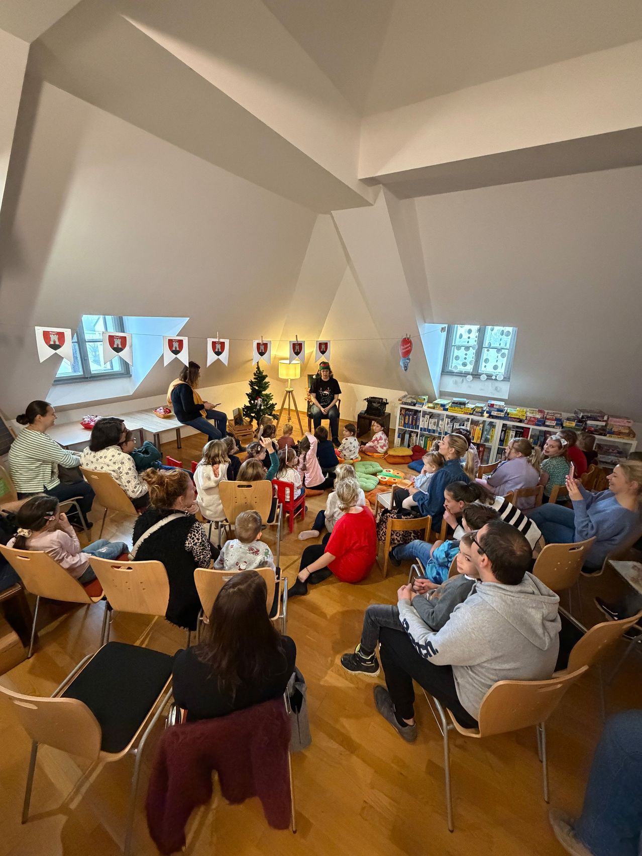 Eine Gruppe von Menschen und Kindern sitzt in Stühlen um einen Raum mit einem Weihnachtsbaum und einem Bücherregal. Eine Frau steht vor einem Schreibtisch.