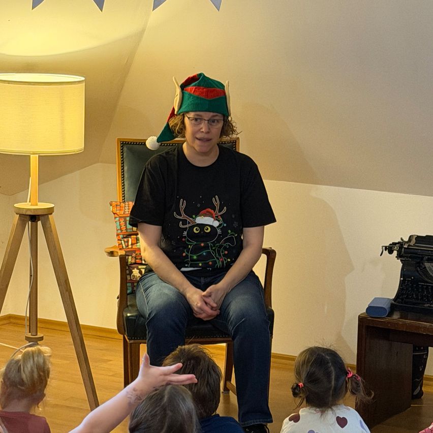 Eine Frau mit einer Weihnachtsmütze sitzt auf einem Stuhl und liest einer Gruppe von Kindern vor. Der Raum ist mit Weihnachtsdekorationen geschmückt.