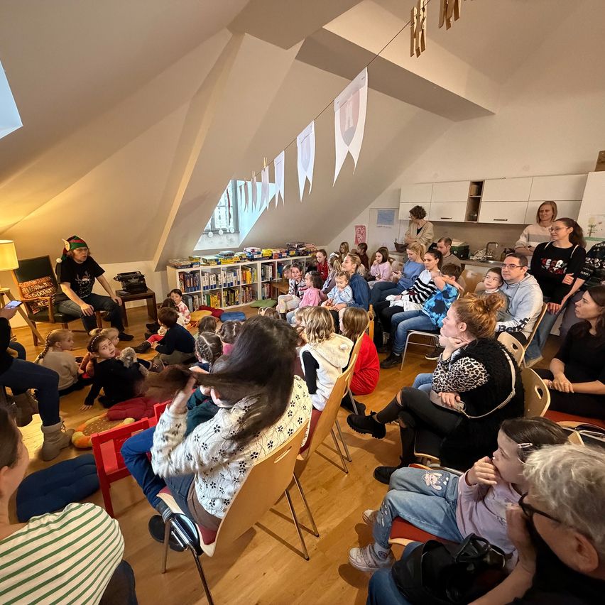 Eine Gruppe von Kindern und Erwachsenen sitzt in einem Dachboden, einige Erwachsene stehen. Ein Mann sitzt auf einem Hocker, und ein Bücherregal ist hinter ihnen. Links steht eine Lampe, und rechts steht ein Schrank.