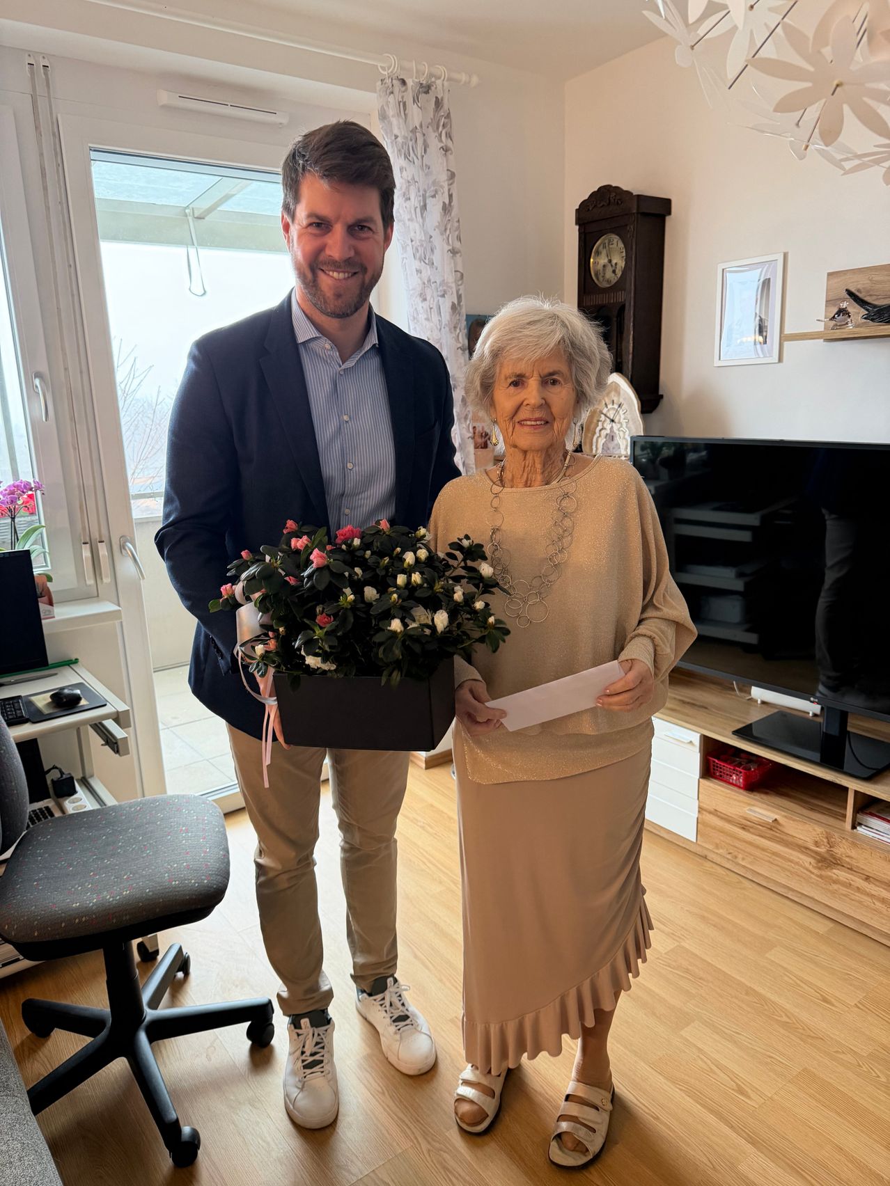 Ein Mann und eine Frau stehen in einem Raum, lächeln und halten einen Blumenstrauß und ein Papier. Ein großes Fenster und eine Uhr sind im Hintergrund.