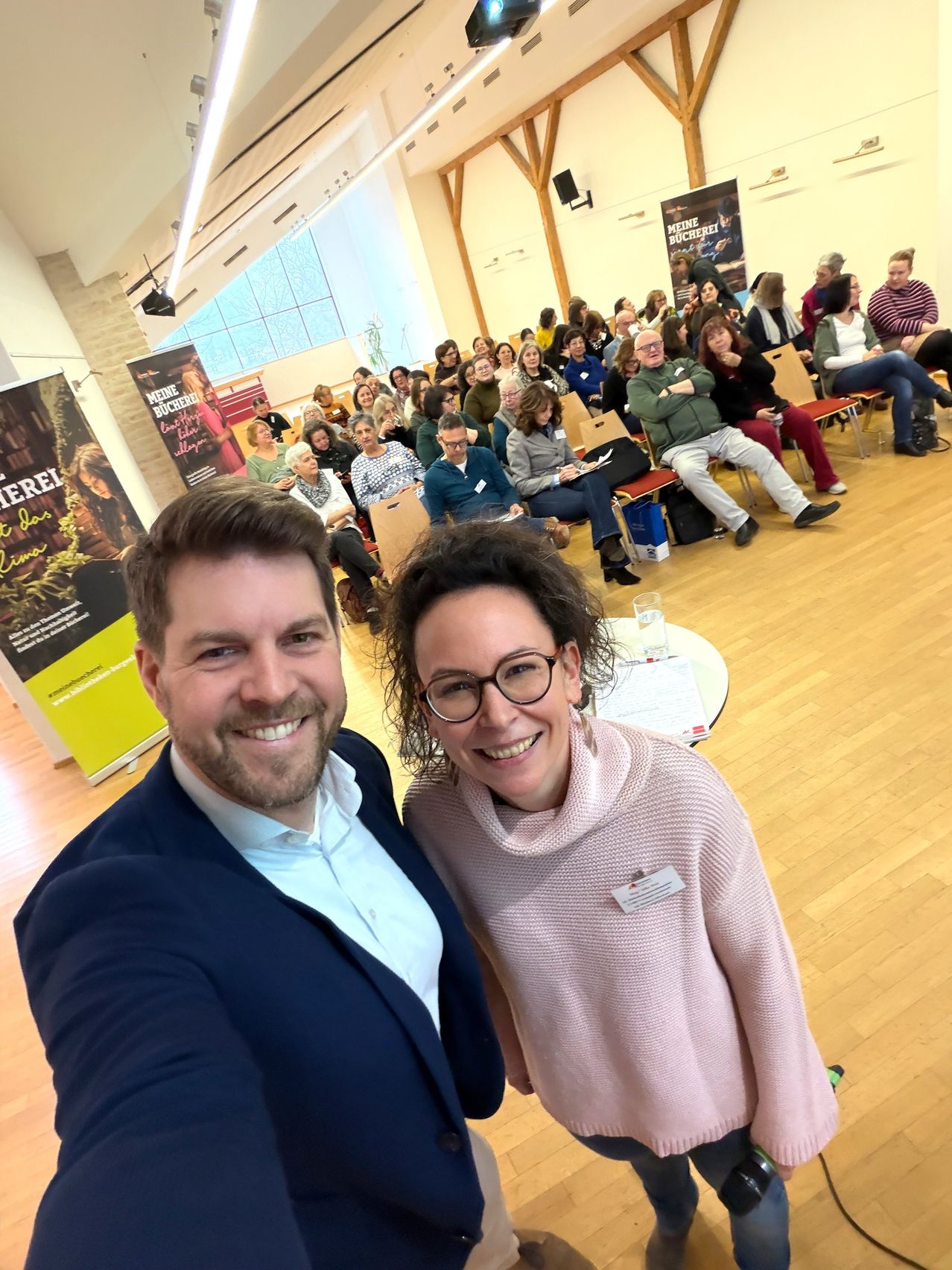 Zwei Personen lächeln und posieren für ein Selfie in einem Raum mit Holzboden. Viele Menschen sitzen vor ihnen auf Stühlen. Einige unterhalten sich. Ein Tisch mit einem Becher und Papieren steht in der Nähe. Banner hängen an der Wand.