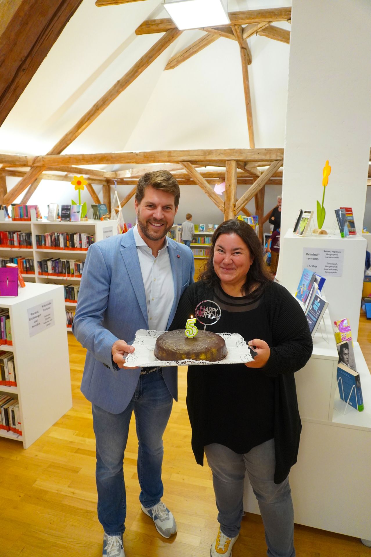 A man and a woman are standing in a library, holding a cake with the number five on it. The woman is smiling and looking at the camera.