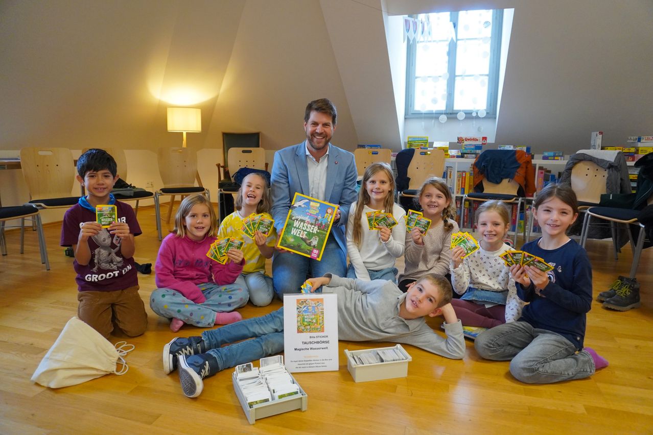 A group of children and a man in a suit sit on the floor holding cards and books. A boy lies on the floor smiling.