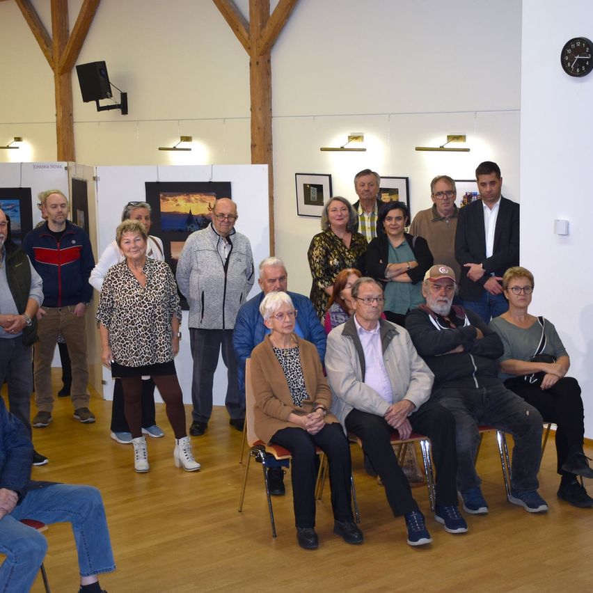 A group of people in an art gallery, some standing and others sitting, surrounded by framed pictures on the wall. A clock is mounted on the wall.