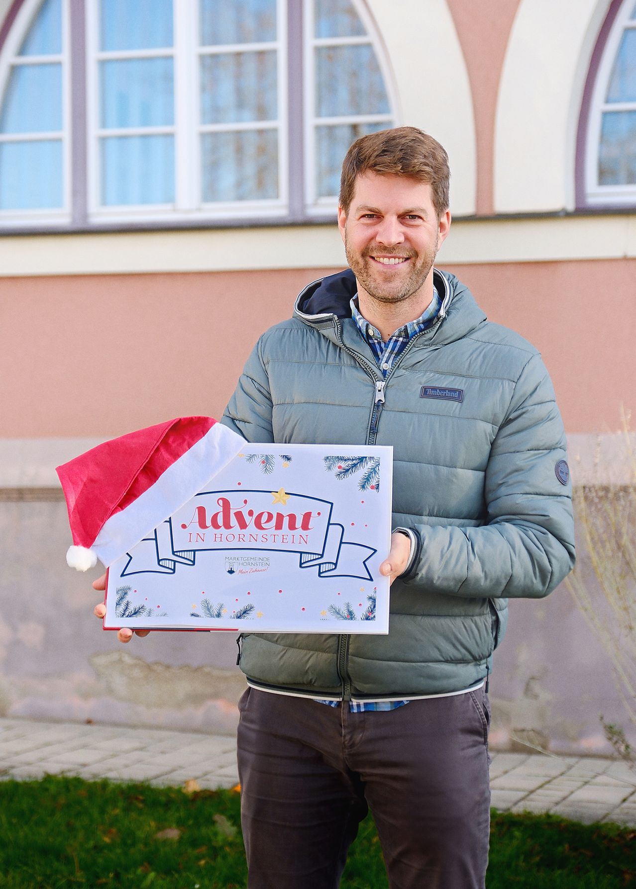 A man in a Timberland jacket is smiling and holding a box that reads Advent in Hornstein. He is wearing a Santa hat and is standing in front of a building.
