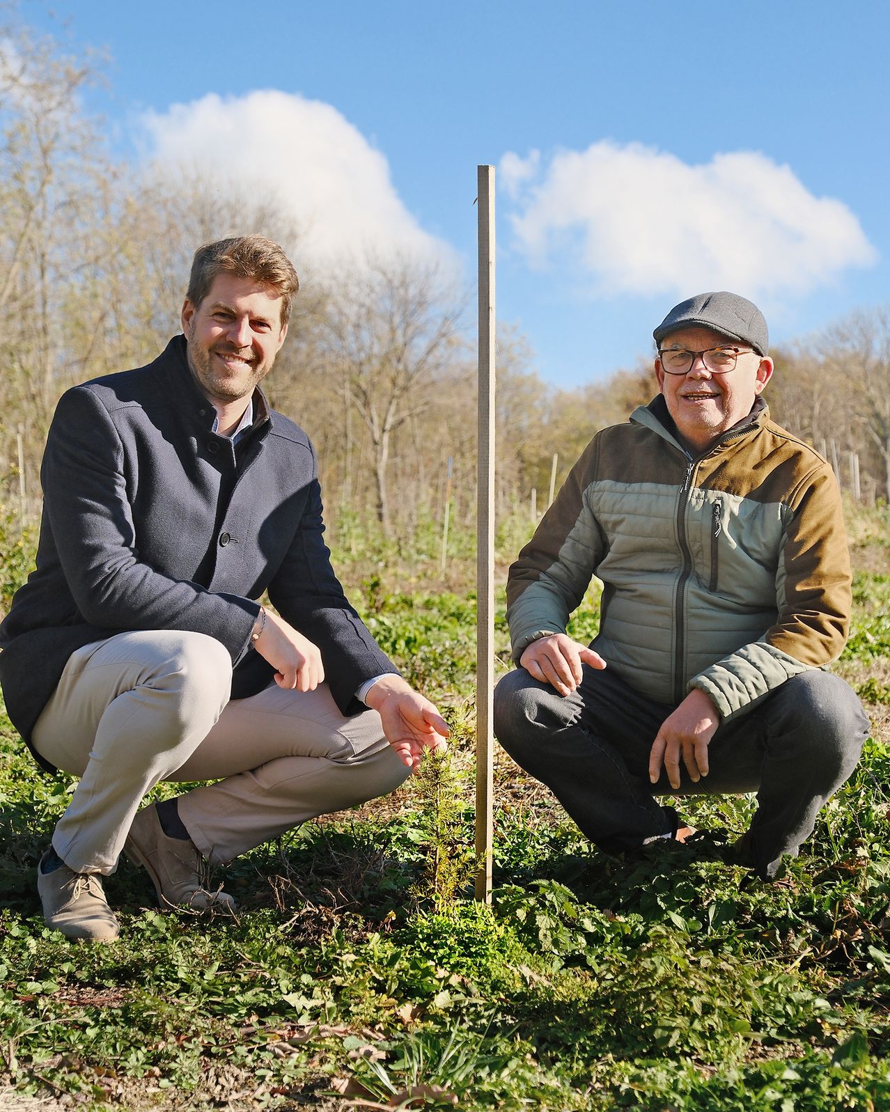 Two men crouch next to a tall wooden pole in a grassy field with trees in the background under a blue sky with clouds.