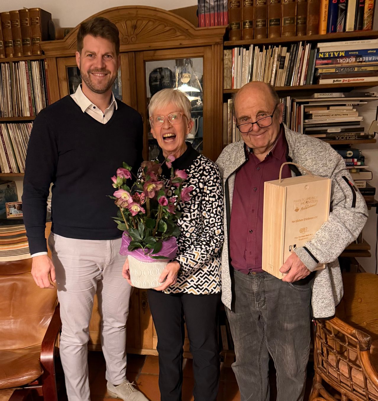 Three people stand in a room with bookshelves behind them. A man on the left smiles, a woman in the middle holds flowers, and a man on the right holds a box.