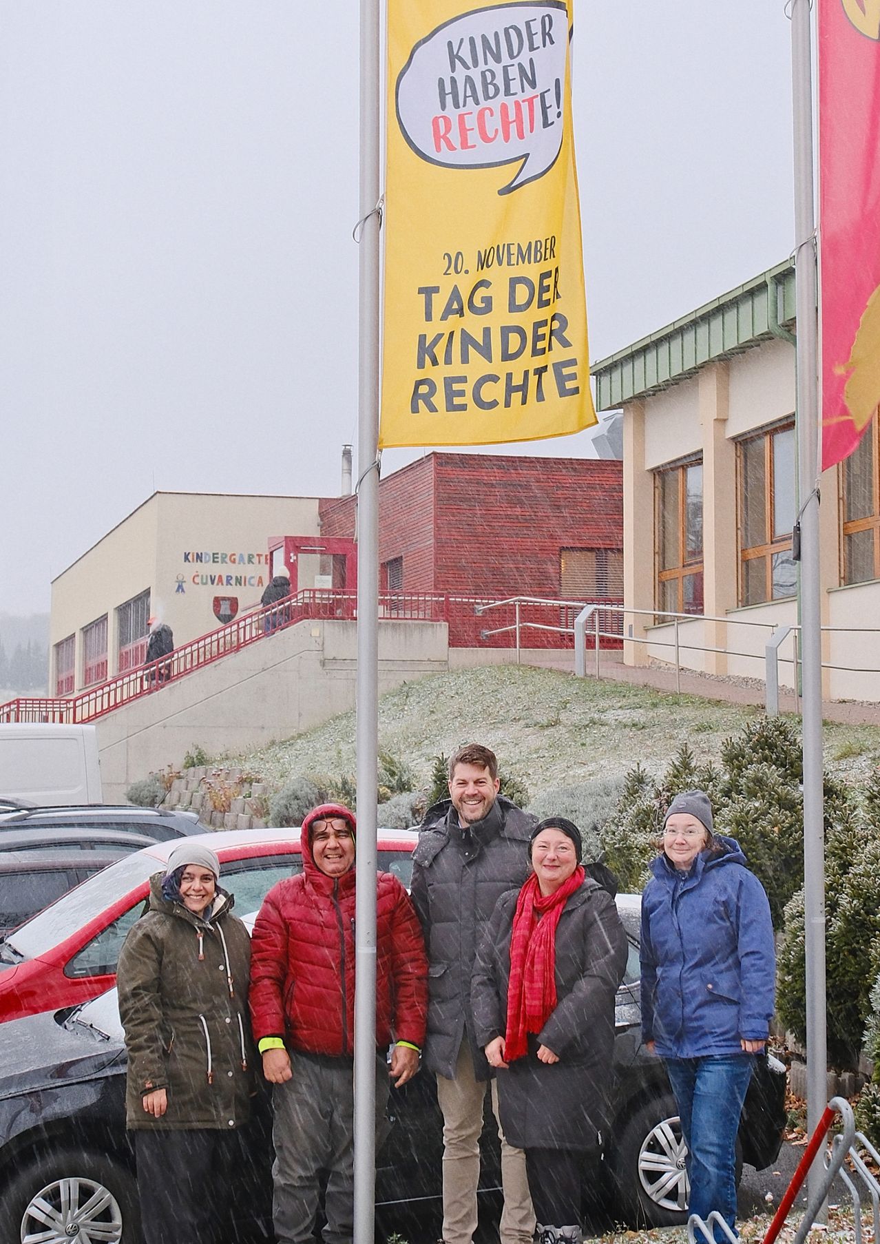 Eine Gruppe von Menschen steht vor einem gelben Banner mit den Worten 'Tag der Kinder Rechte' und einem Gebäude mit einer roten Treppe.