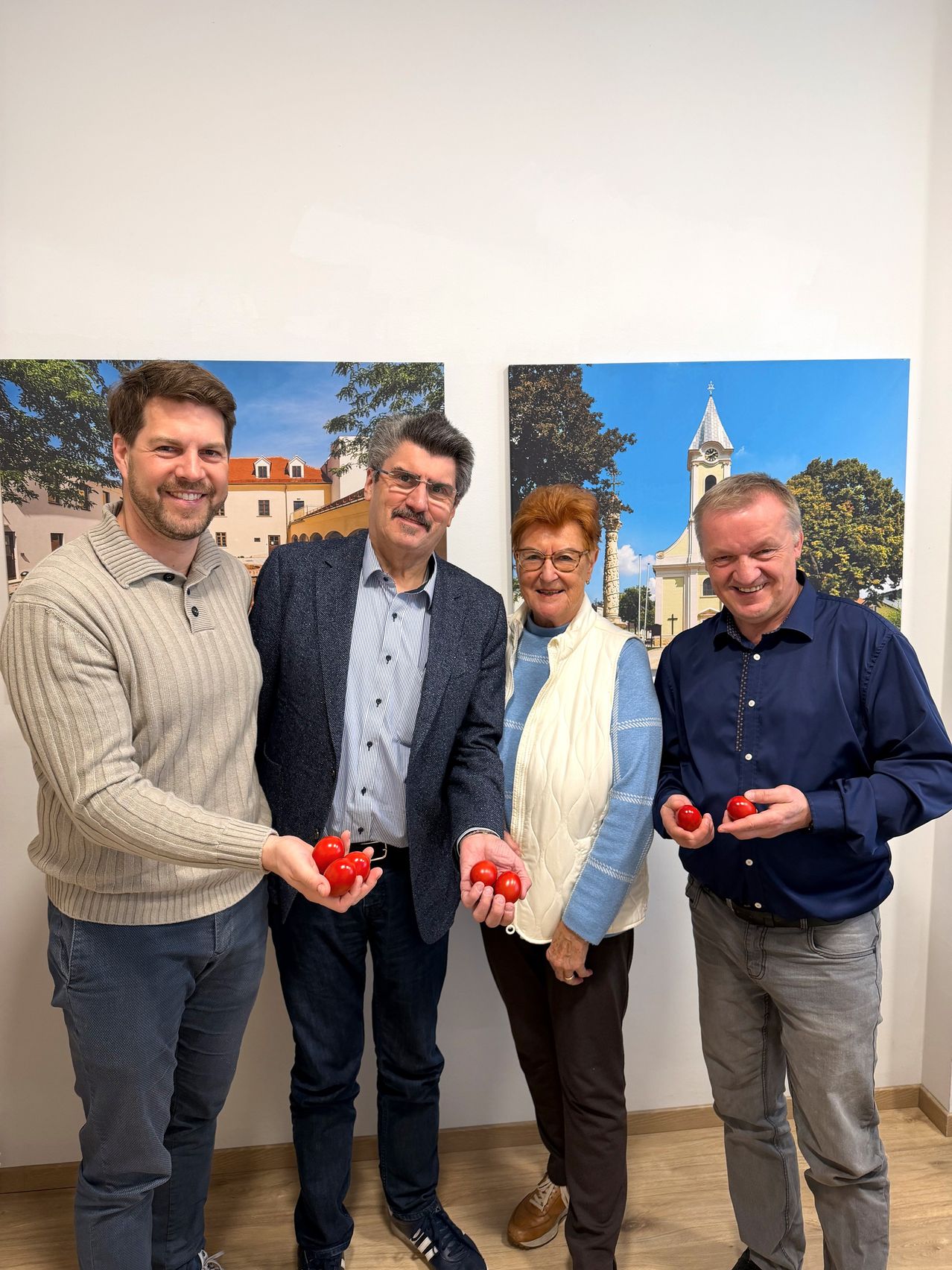 Four people stand together smiling, with one person holding red apples. Behind them are two large paintings.