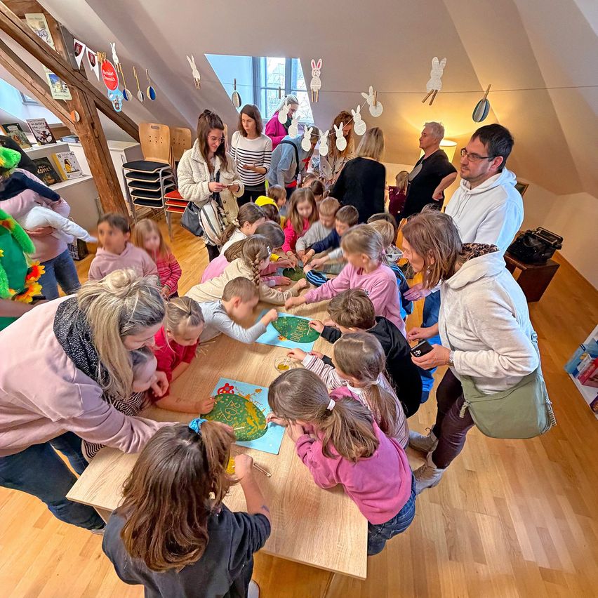 Several adults and children gather around a table in a room with wooden floors, engaged in an art activity. Some children are painting, and others are watching. Easter decorations hang from the ceiling.