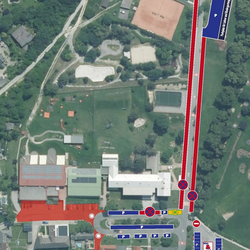 An aerial view of a campus with multiple buildings, parking areas, and pathways. The main entrance has a blue sign with 'P' and 'H' markings. A red line indicates a bus route.